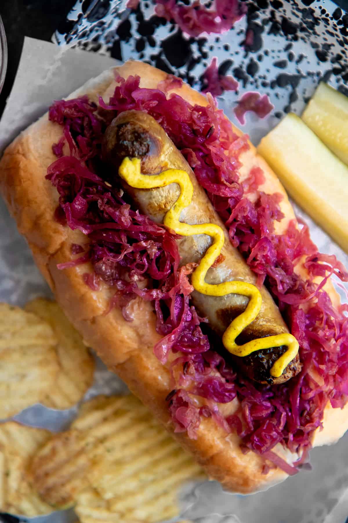 A brat drizzled with mustard sits on a bed of red sauerkraut on top of a bun. Potato chips and a sliced pickle surround the brat.