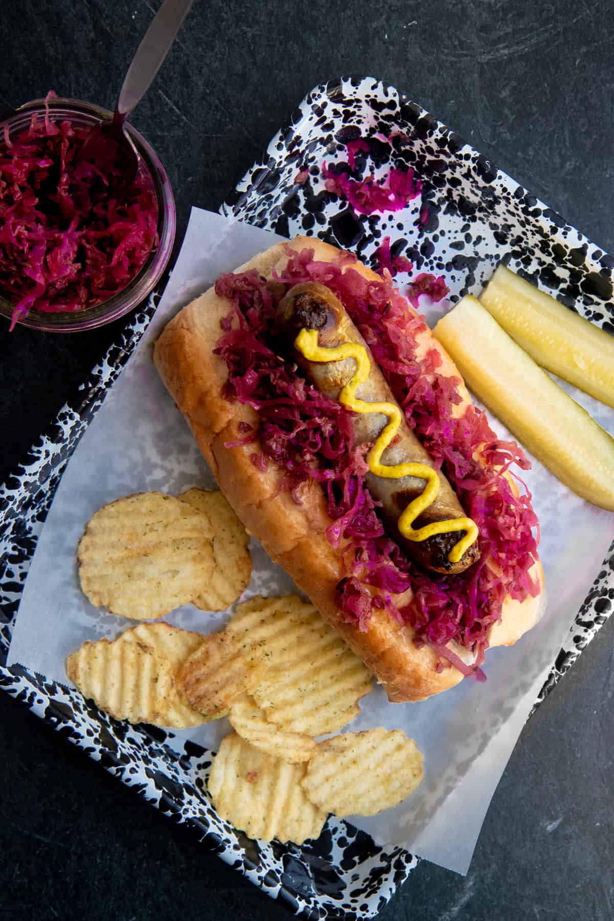 A brat drizzled with mustard sits on a bed of homemade sauerkraut on top of a bun. Potato chips and a sliced pickle surround the brat.