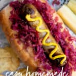 A brat drizzled with mustard sits on a bed of red sauerkraut on top of a bun. Potato chips and a sliced pickle surround the brat. A text overlay reads "Easy Homemade Sauerkraut."