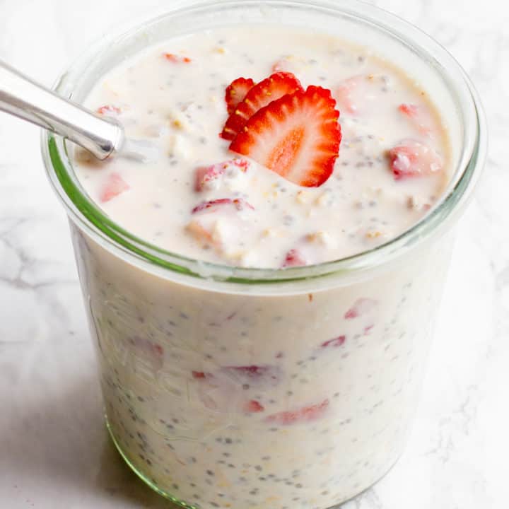 A glass jar filled with strawberry cheesecake overnight oats sits on a marble countertop. The oats are garnished with strawberry slices, and a spoon dips into the jar.