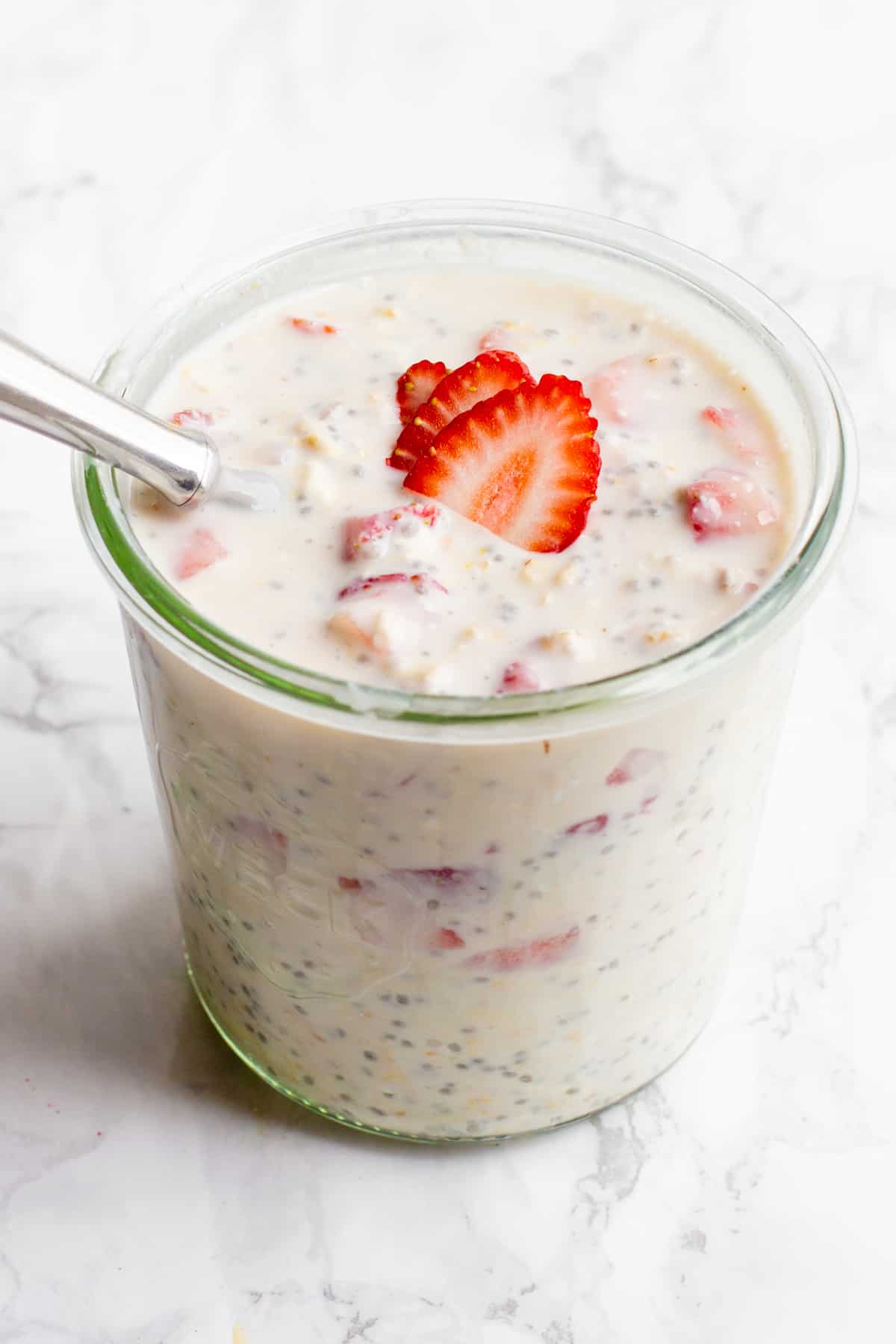 A glass jar filled with strawberry cheesecake overnight oats sits on a marble countertop. The oats are garnished with strawberry slices, and a spoon dips into the jar.