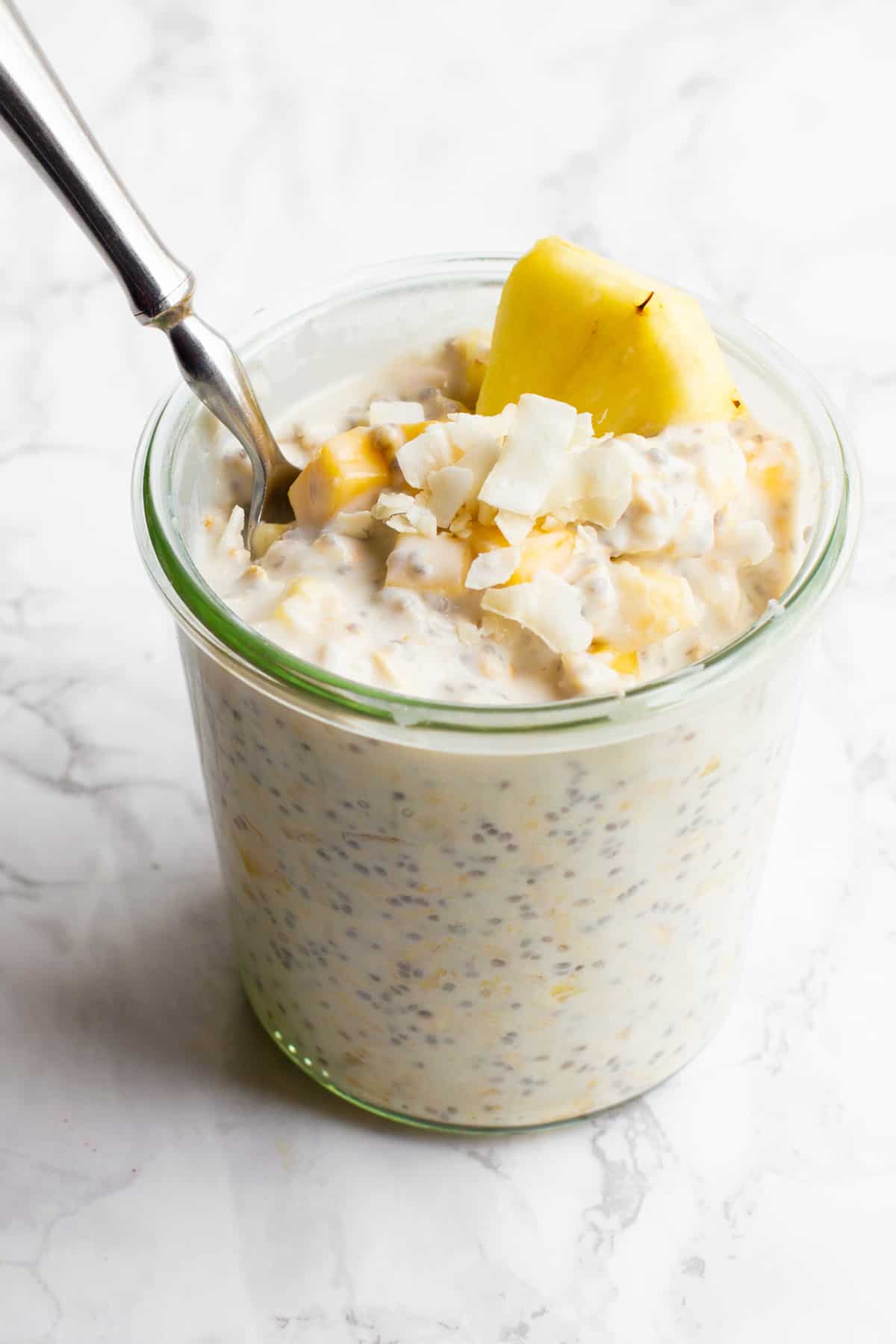 A glass jar filled with tropical coconut overnight oats sits on a marble countertop. The oats are garnished with pineapple and coconut, and a spoon dips into the jar.