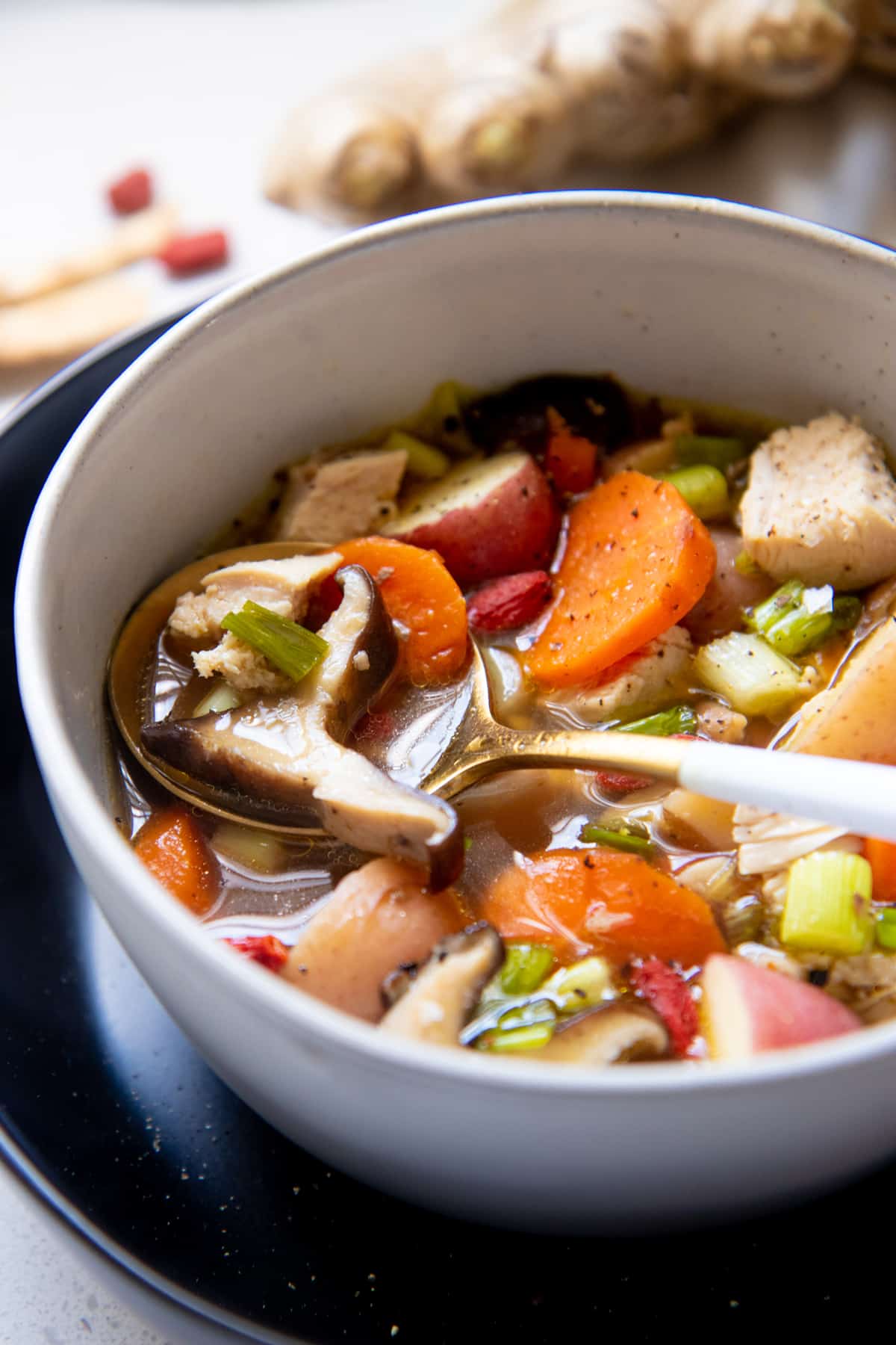 A spoon dips into a bowl full of soup broth, chicken, vegetables, and Chinese herbs.