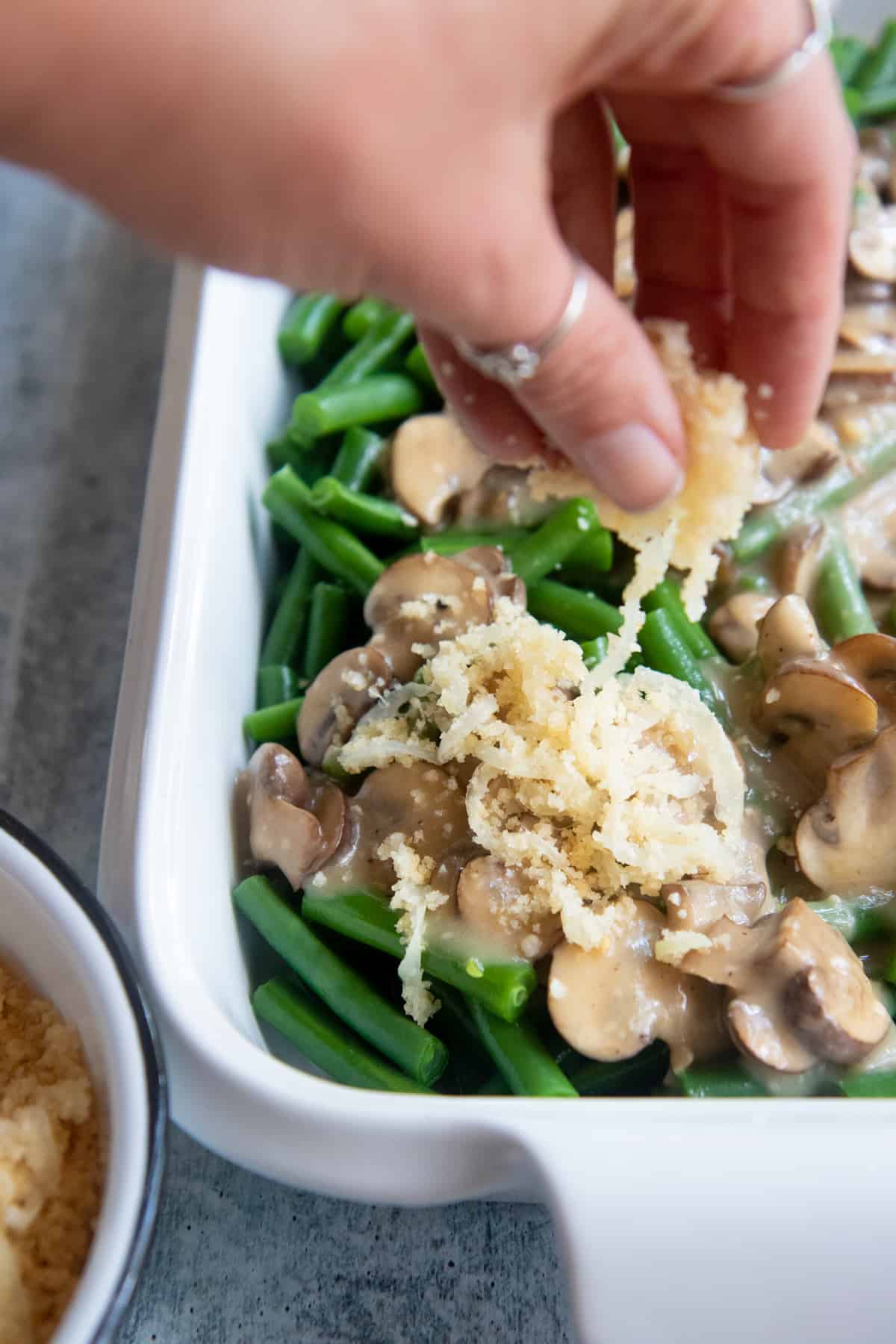 A hand sprinkles Crispy Parmesan Onions over the top of a baking dish of green beans and mushroom sauce.