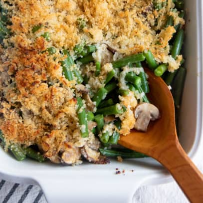 A wooden spoon scoops some fresh green bean casserole out of a white baking dish.