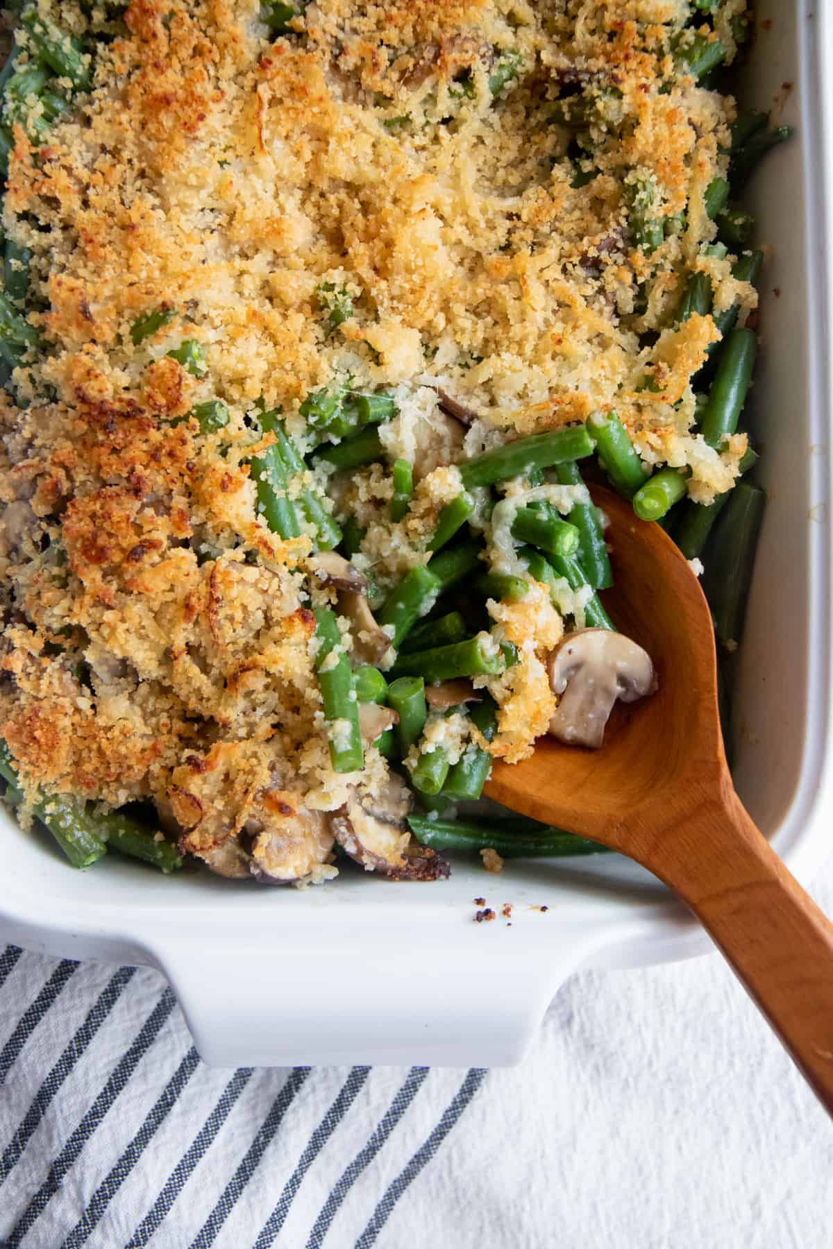 A wooden spoon scoops some fresh green bean casserole out of a white baking dish.