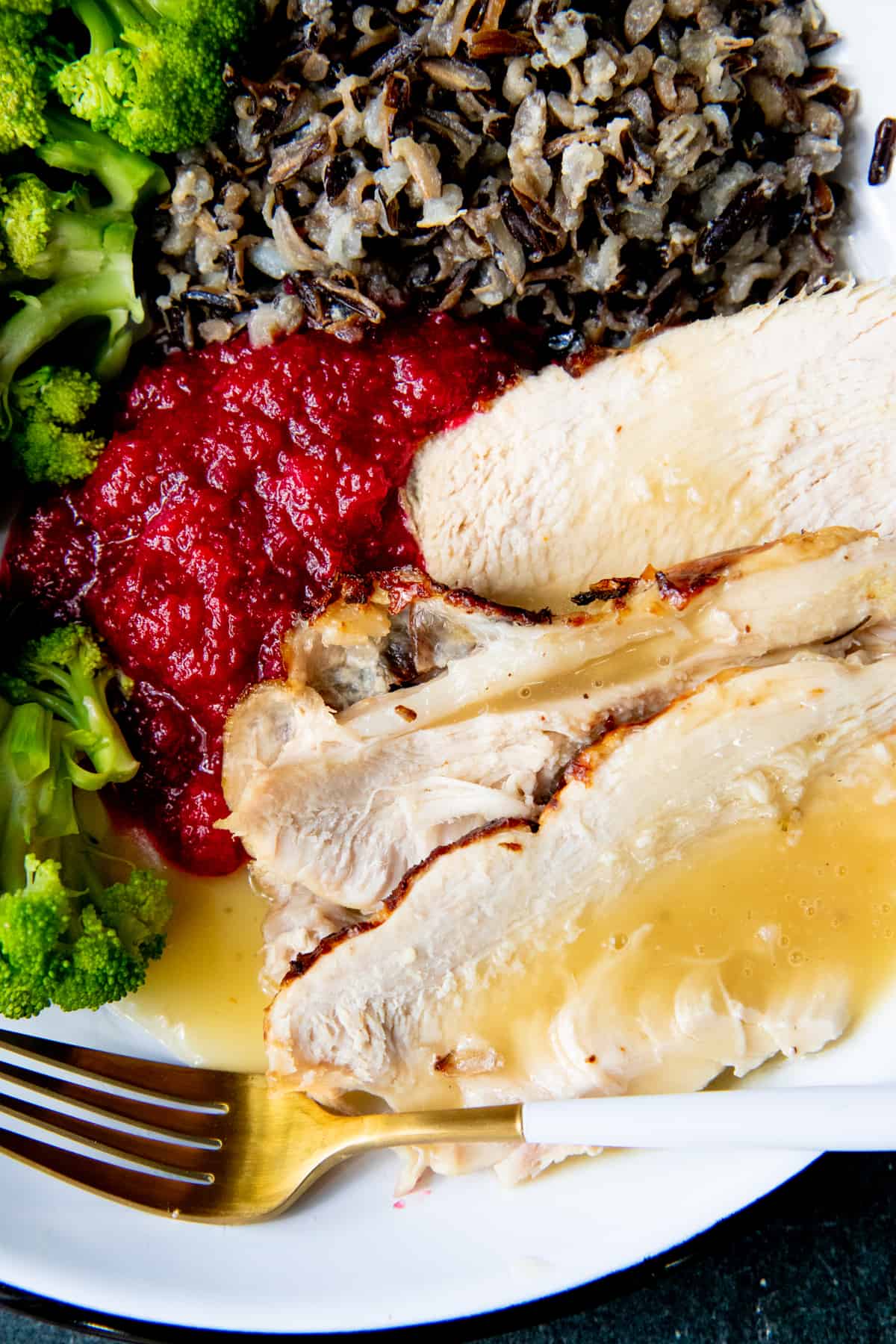Slices of turkey covered in gravy, next to piles of Thanksgiving side dishes, such as broccoli and cranberry applesauce.
