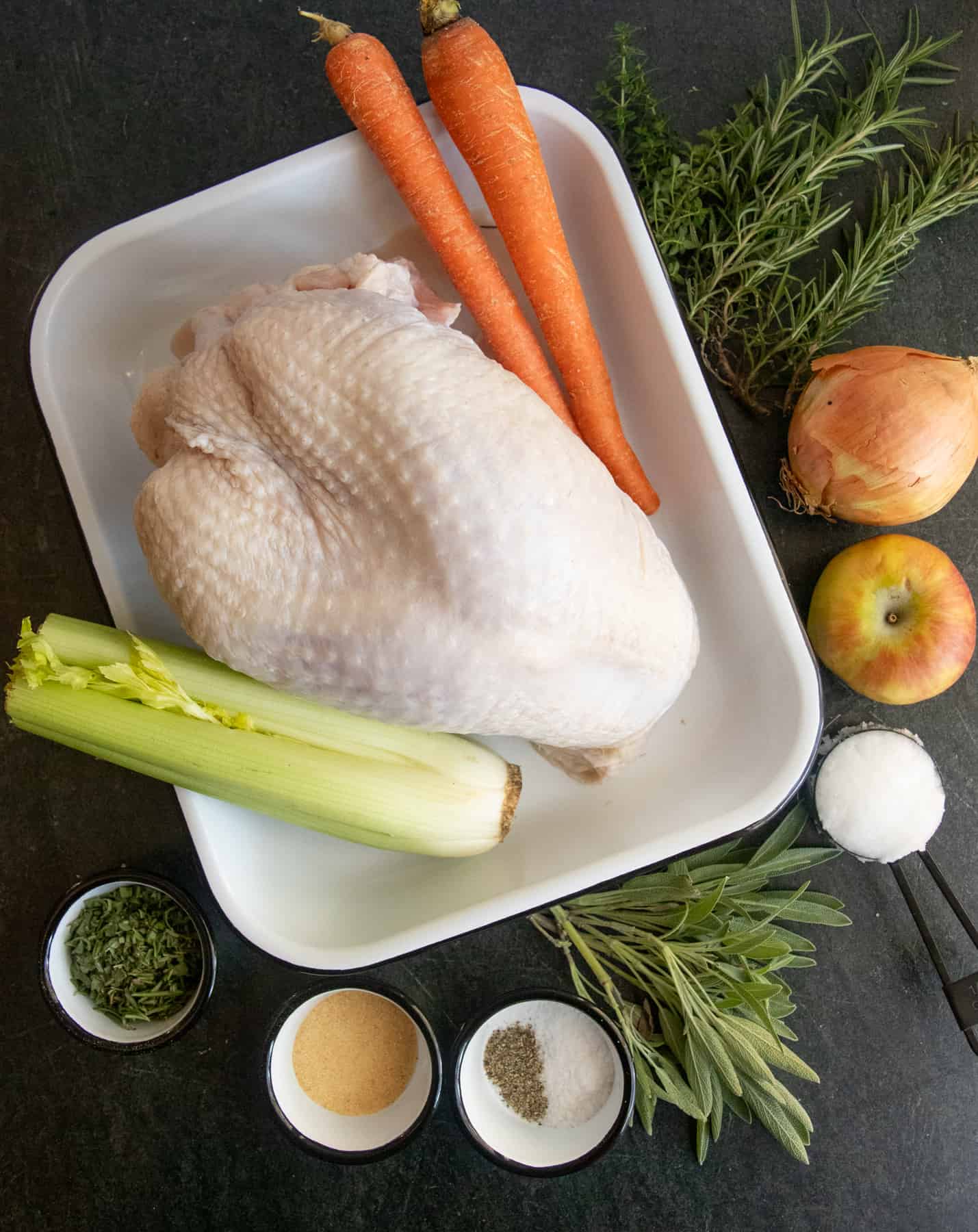 Ingredients sit on a white plate on a dark background - turkey breast, carrots, celery, apple, onion, herbs, and spices.