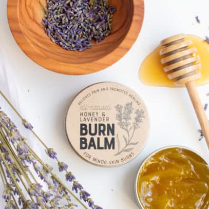 An open tin of burn balm sits in front of a lidded and labeled tin. A honey dipper, bowl of lavender buds, and sprigs of lavender are nearby.