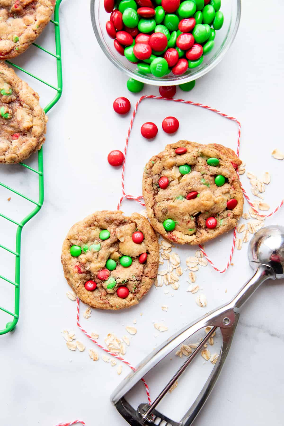 Two cookies dotted with holiday colored M&Ms sit on a white tabletop, surrounded by baker's twine, M&Ms, and rolled oats. A cookie scoop lies next to the cookies.