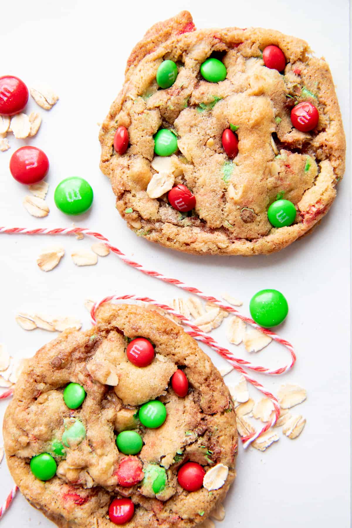 Two cookies dotted with holiday colored M&Ms sit on a white tabletop, surrounded by baker's twine, M&Ms, and rolled oats.