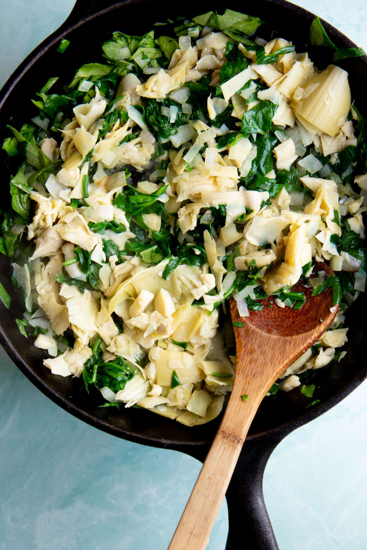 A wooden spoon stirs artichoke hearts with spinach in a skillet.