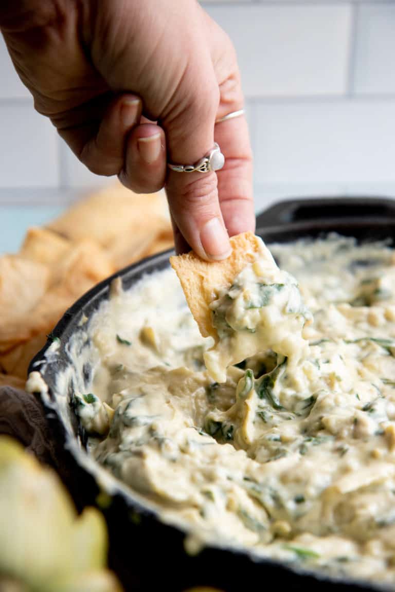 A hand dips a pita chip into a skillet of vegan spinach artichoke dip.