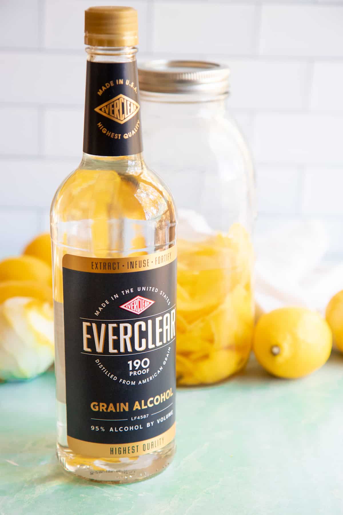 Bottle of Everclear sitting in front of a pile of lemons and a mason jar filled with Everclear and lemon peels.
