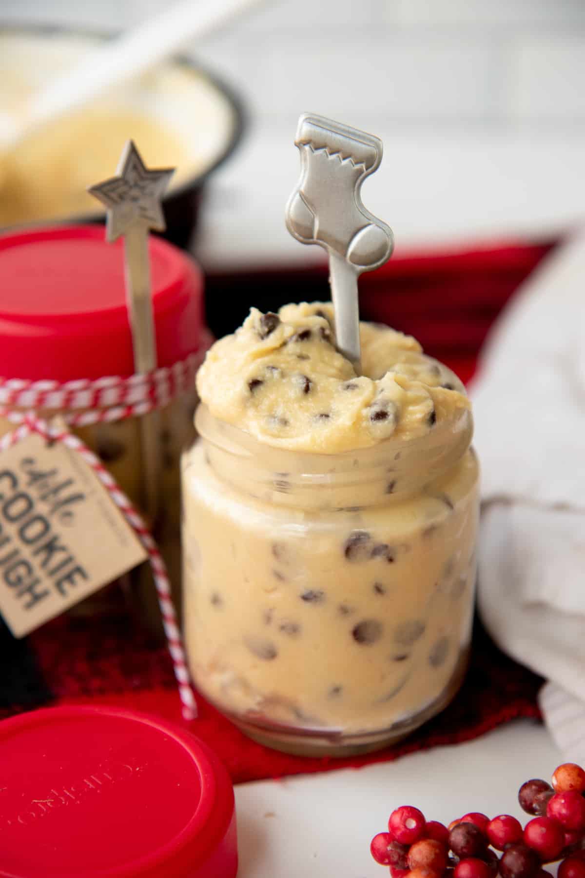 An open jar of eggless cookie dough with a silver holiday spoon in it sits in front of a packaged jar of the dough.