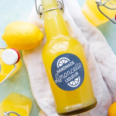 A full and labeled bottle of Homemade Limoncello lies on a white kitchen towel among lemons and other filled bottles.