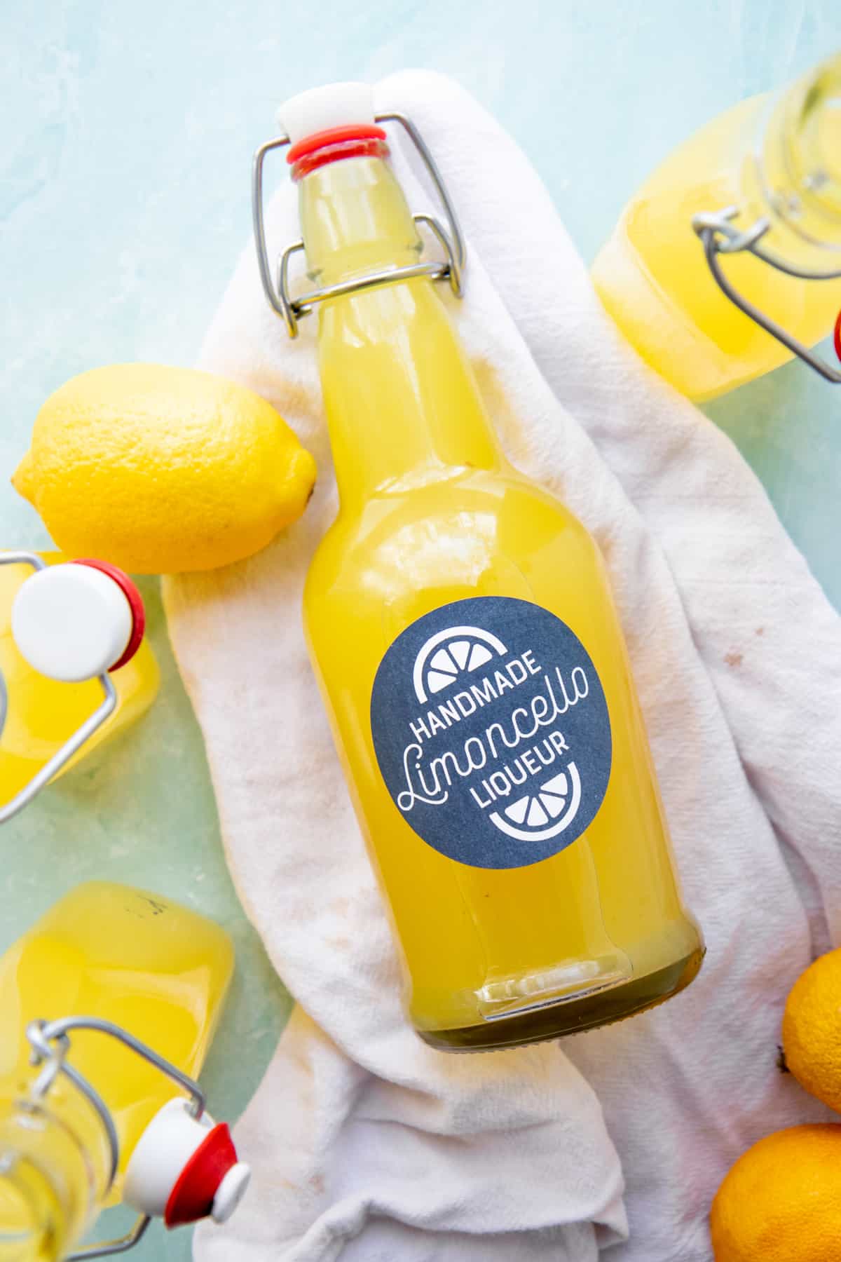 A full and labeled bottle of Homemade Limoncello lies on a white kitchen towel among lemons and other filled bottles.