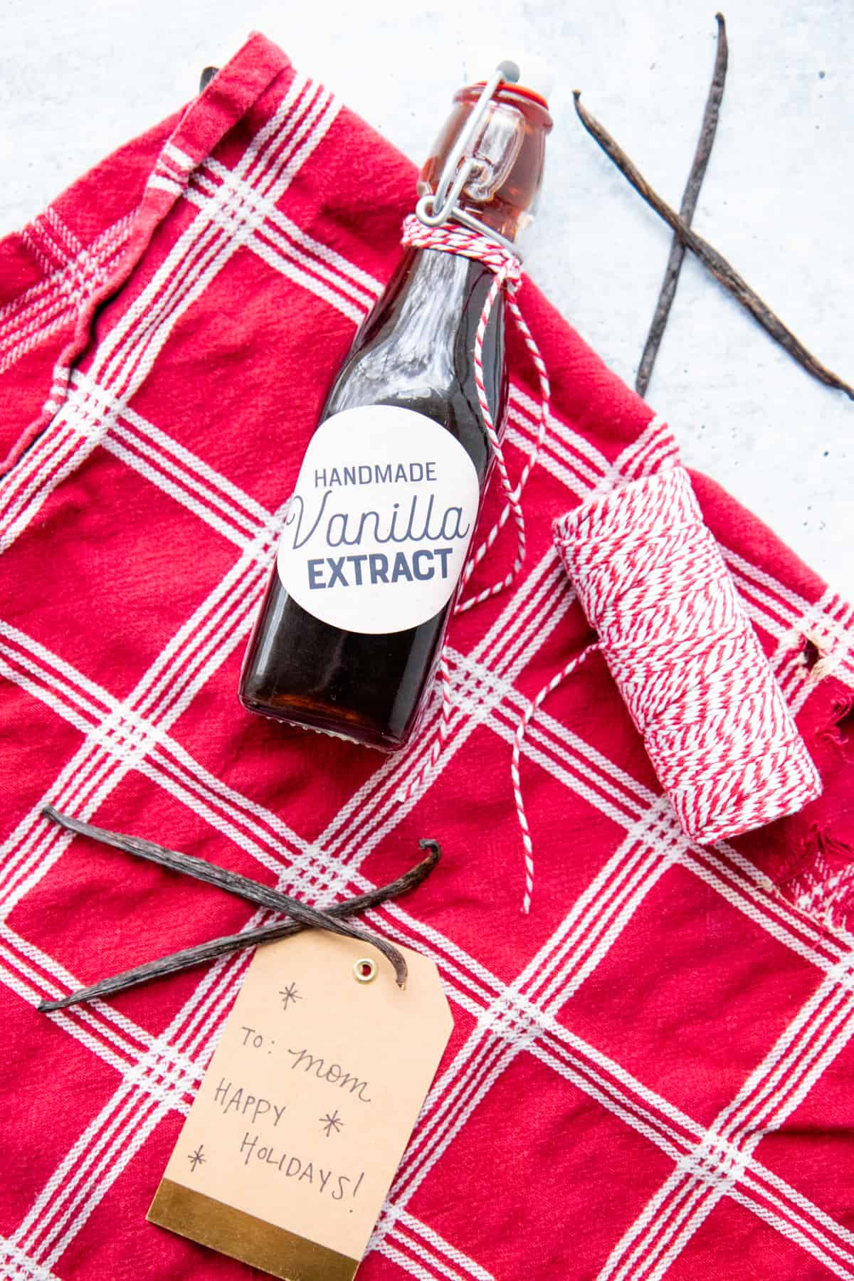 A bottle of homemade vanilla extract lays on red and white fabric, surrounded by whole vanilla beans.