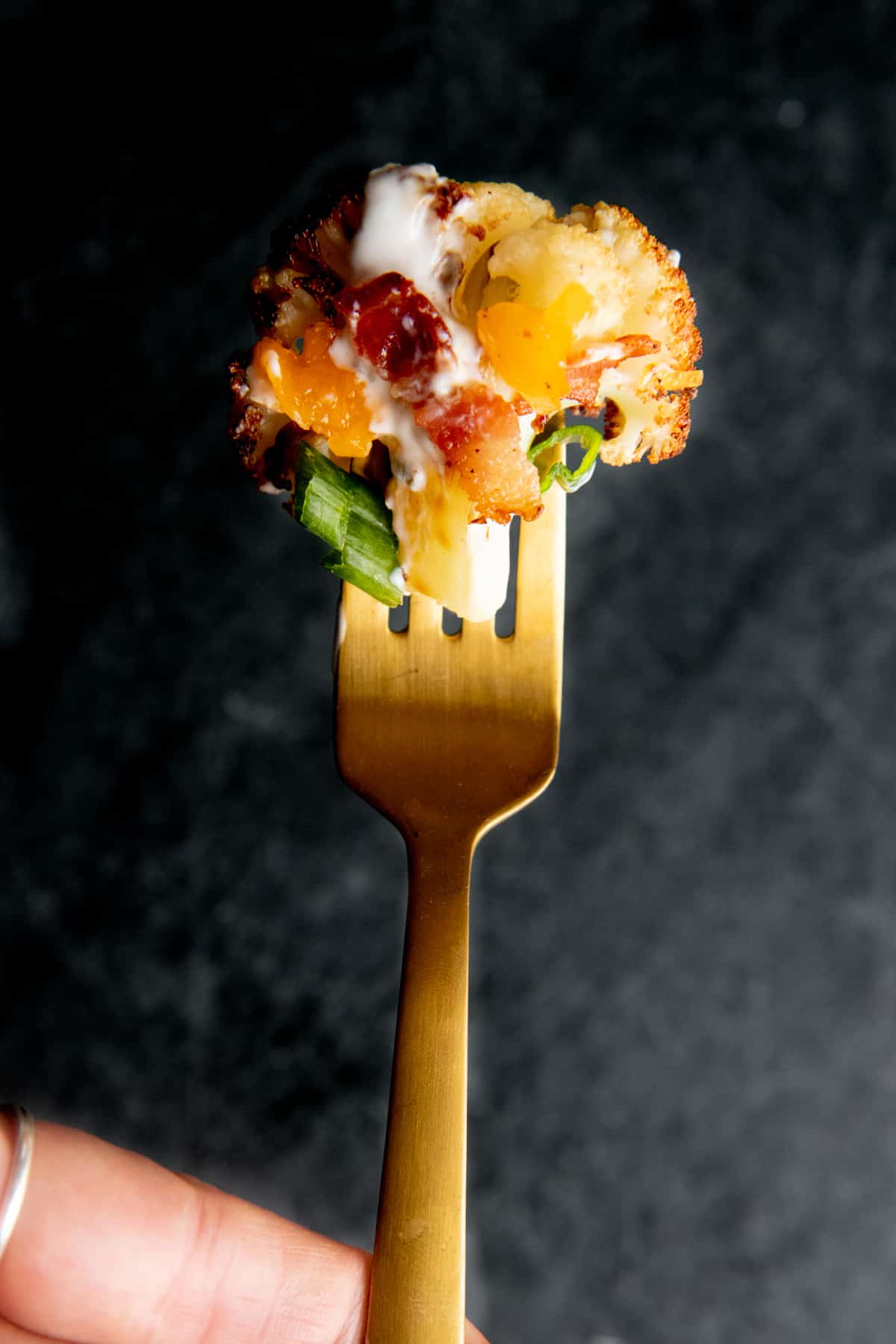A gold fork holds a bite of loaded roasted cauliflower in front of a dark background.