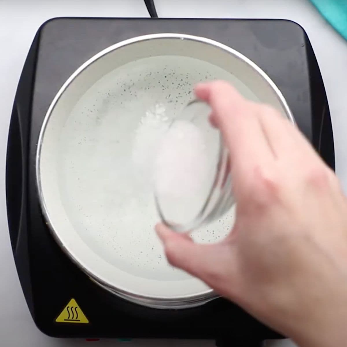 A hand pours salt into boiling water in a pot on a black hot plate
