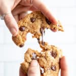 Two hands pull an almond flour chocolate chip cookie apart. Gooey chocolate stretches between the two cookie halves.