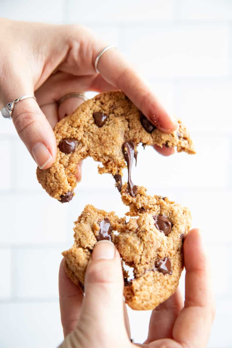 Two hands pull an almond flour chocolate chip cookie apart. Gooey chocolate stretches between the two cookie halves.