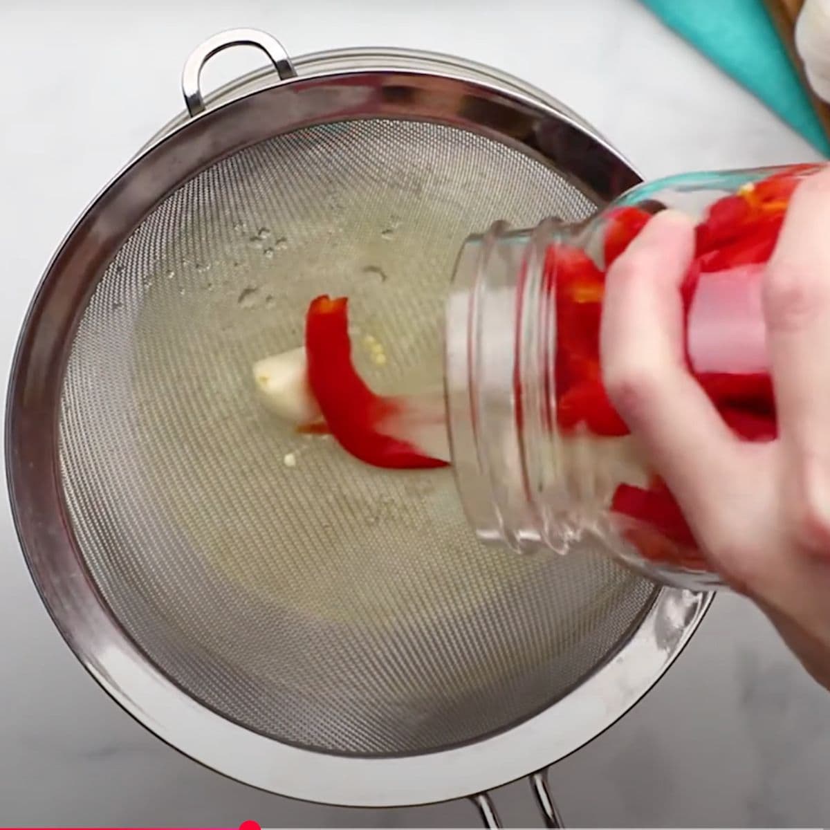 A hand pours out a jar of fermented peppers and garlic into a metal sieve