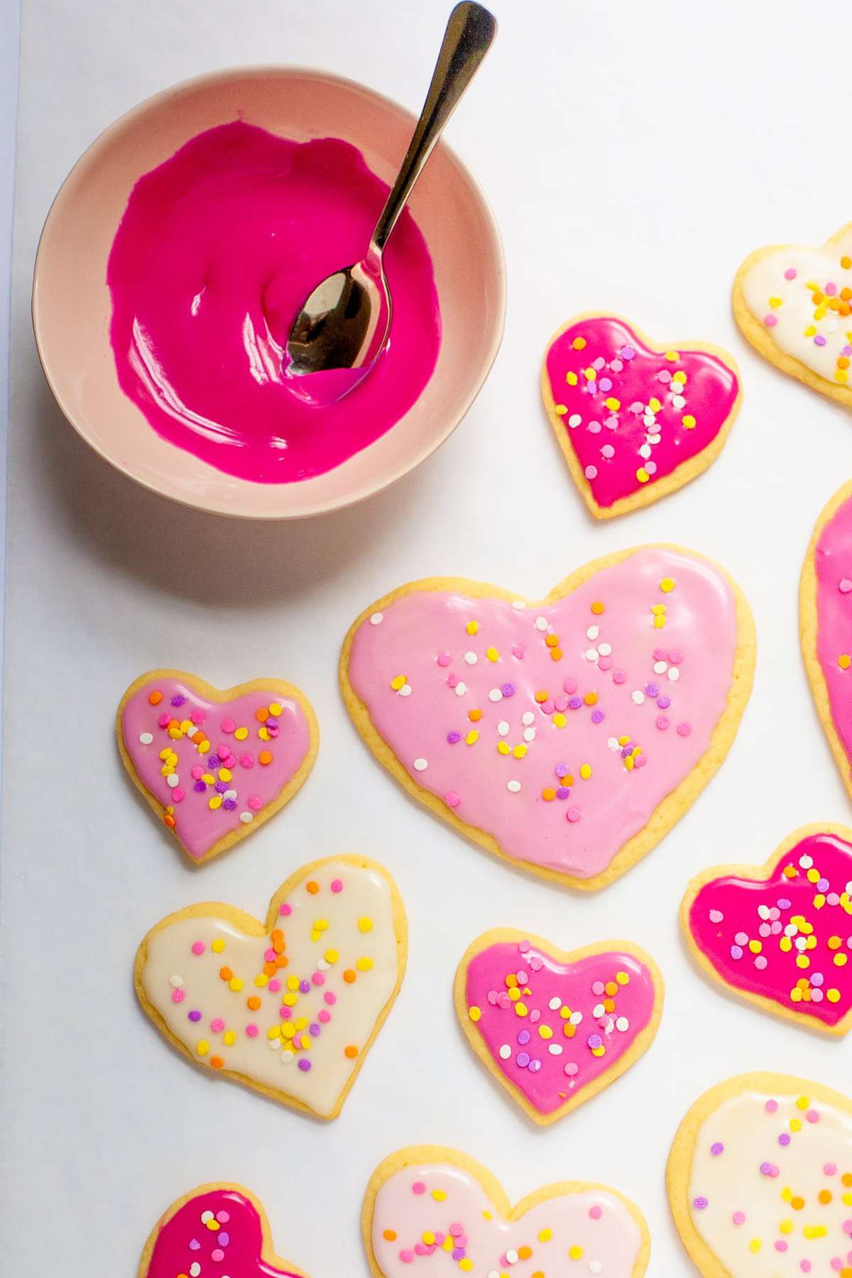 A pink bowl of sugar cookie icing sits next to heart-shaped cookies.