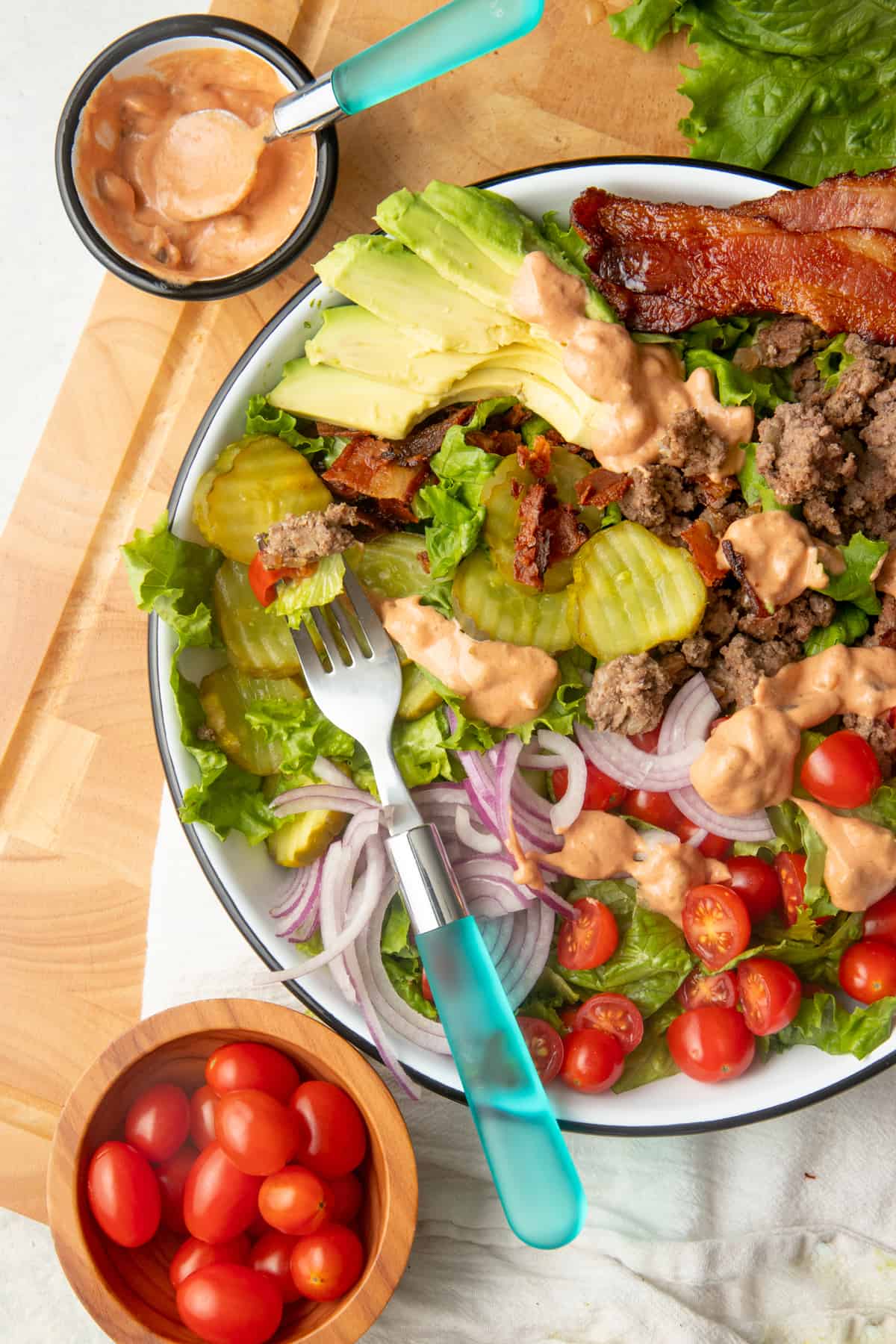A fork with a blue handle rests in a large bowl filled with burger salad ingredients.