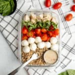 A glass dish filled with grain bowl ingredients sits on a brown and white dishtowel. Tomatoes and mozzarella sit on the side.