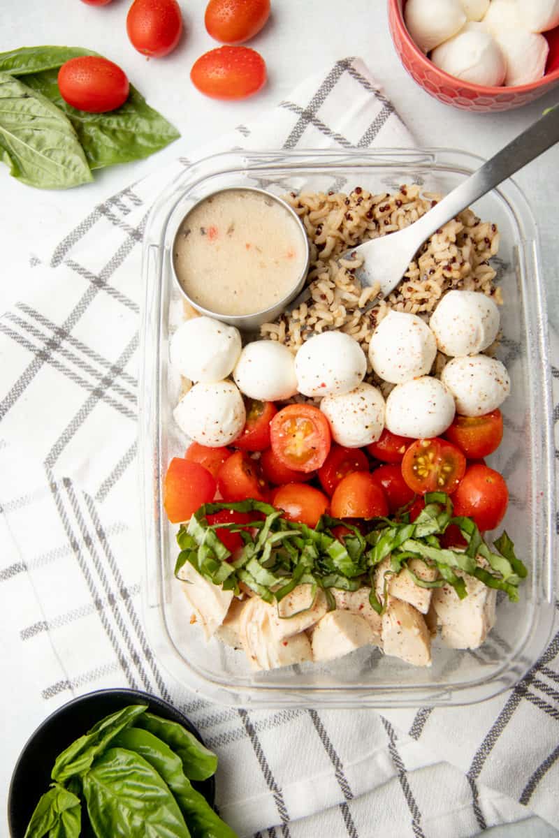 A glass dish is arranged with ingredients for caprese grain bowl. A cup of dressing rests in the upper left corner.