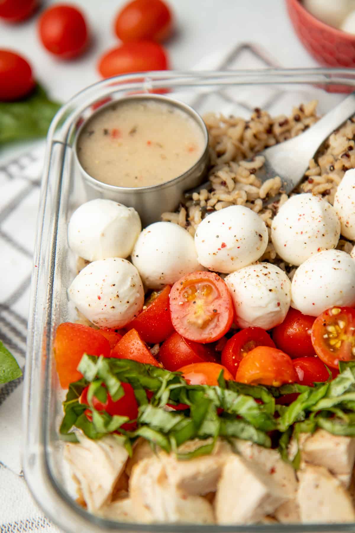 A glass dish is arranged with ingredients for caprese grain bowl. A cup of dressing rests in the upper left corner.