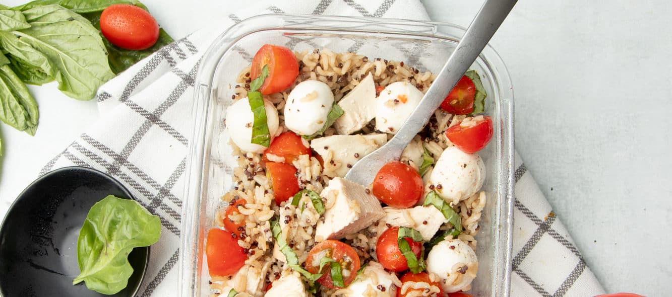 Tomatoes, mozzarella balls, basil, and grains are combined in a rectangular glass container. Container and ingredients sit on a brown and white plaid dishtowel.