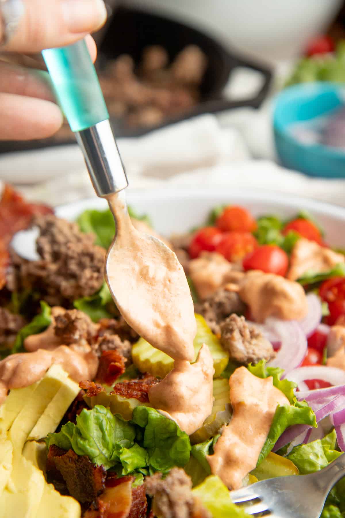 A spoon with a blue handle drizzles "special sauce" over a burger bowl.