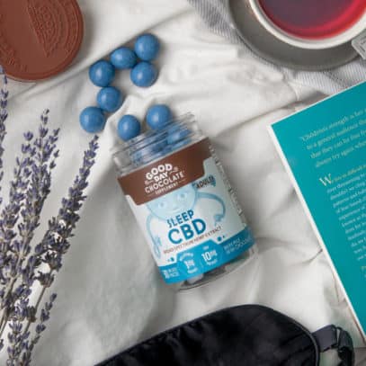 A bottle of Good Day Chocolates "Sleep CBD" spills out onto a white piece of fabric. The bottle is surrounded by a book, sprigs of lavender, a sleep mask, and a cup of tea.