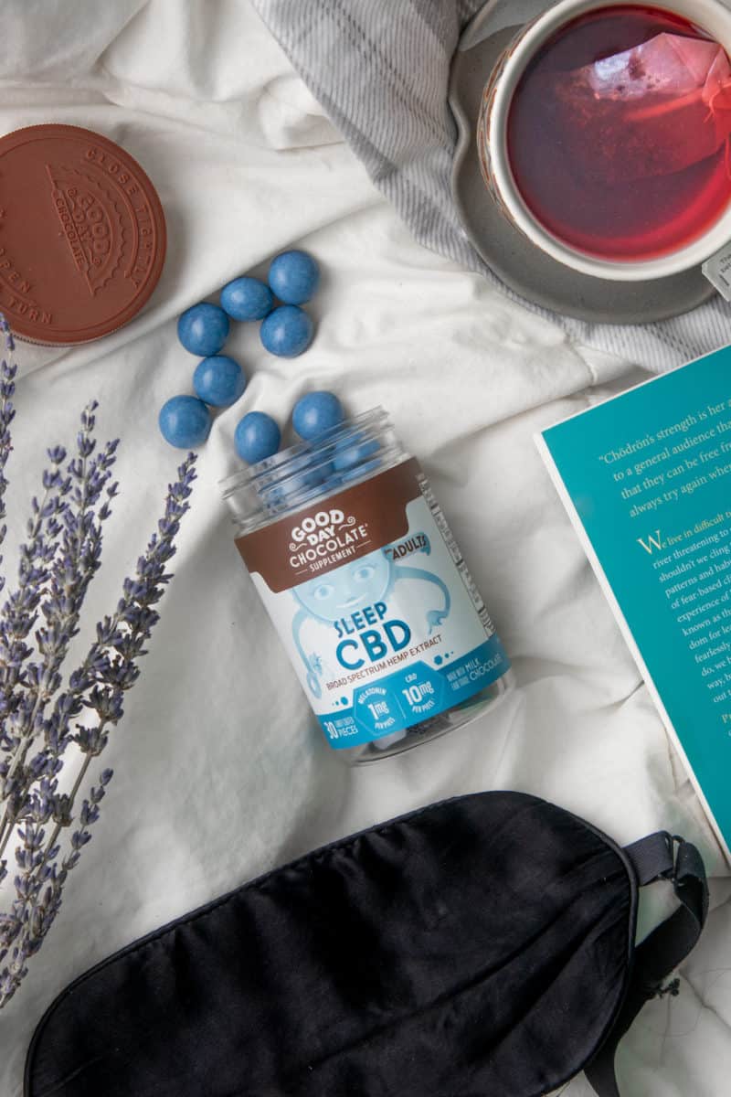 A bottle of Good Day Chocolates "Sleep CBD" spills out onto a white piece of fabric. The bottle is surrounded by a book, sprigs of lavender, a sleep mask, and a cup of tea.