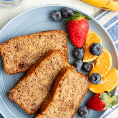 Three slices of almond flour banana bread on a blue plate with mixed fruit, next to a cup of coffee.