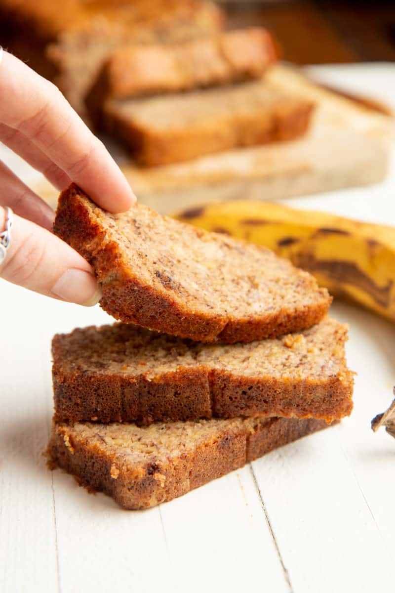 A hand lifts a slice of banana bread from a stack of slices.