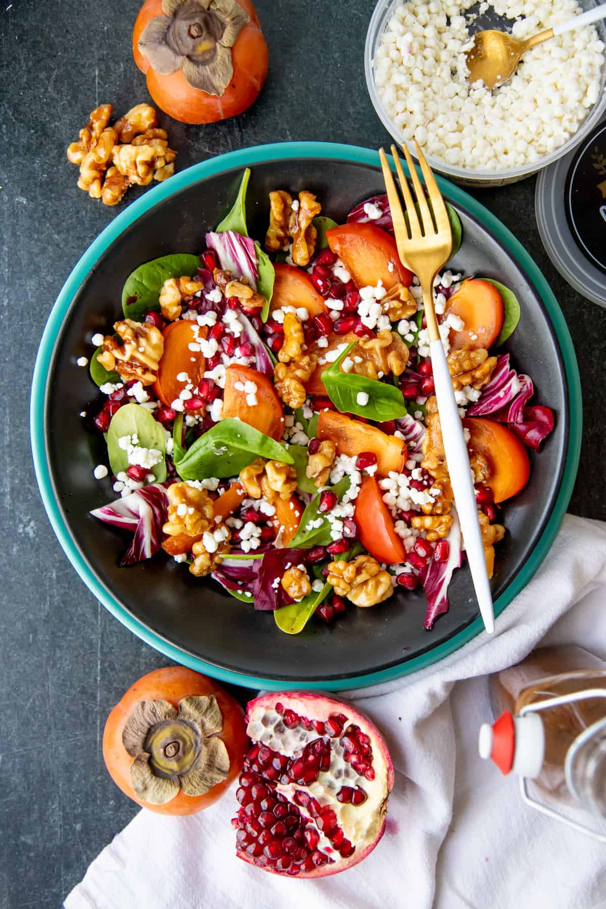 A fork rests on a dark bowl, filled with persimmon and pomegranate salad.