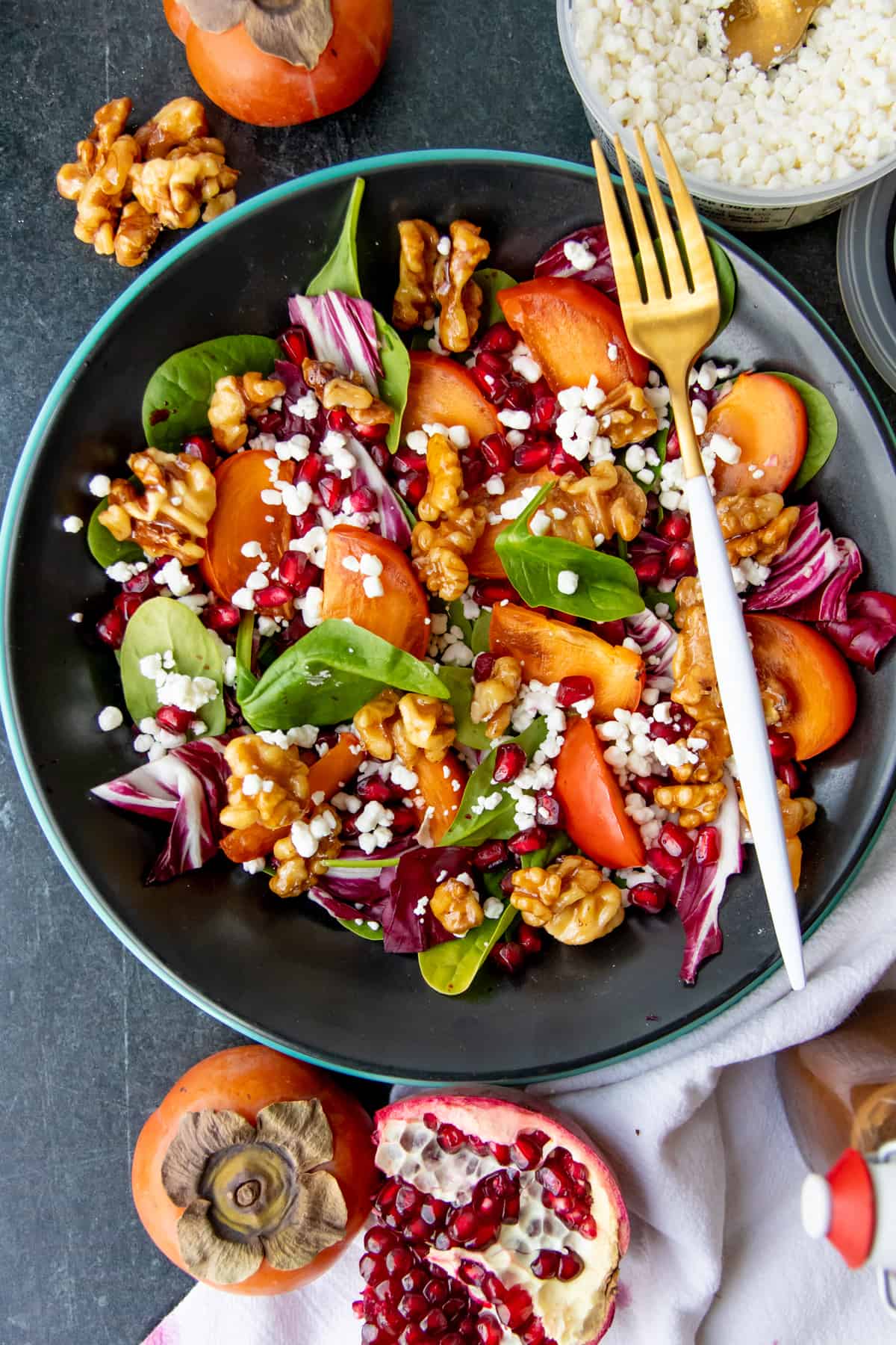 A fork rests on a dark bowl, filled with pomegranate and persimmon salad.