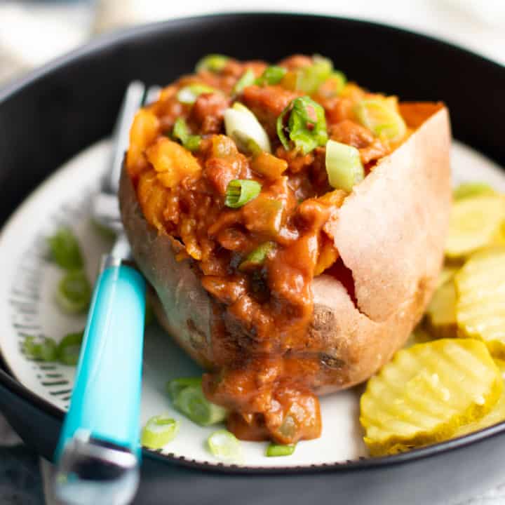 Sweet potato stuffed with Sloppy Joes filling on a plate, with a teal-handled fork and pickle slices next to the potato.