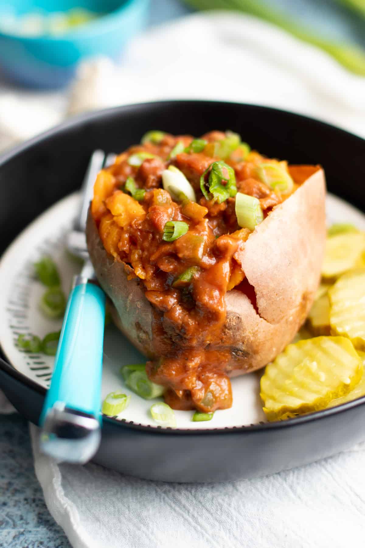 Sweet potato stuffed with Sloppy Joes filling on a plate, with a teal-handled fork and pickle slices next to the potato.