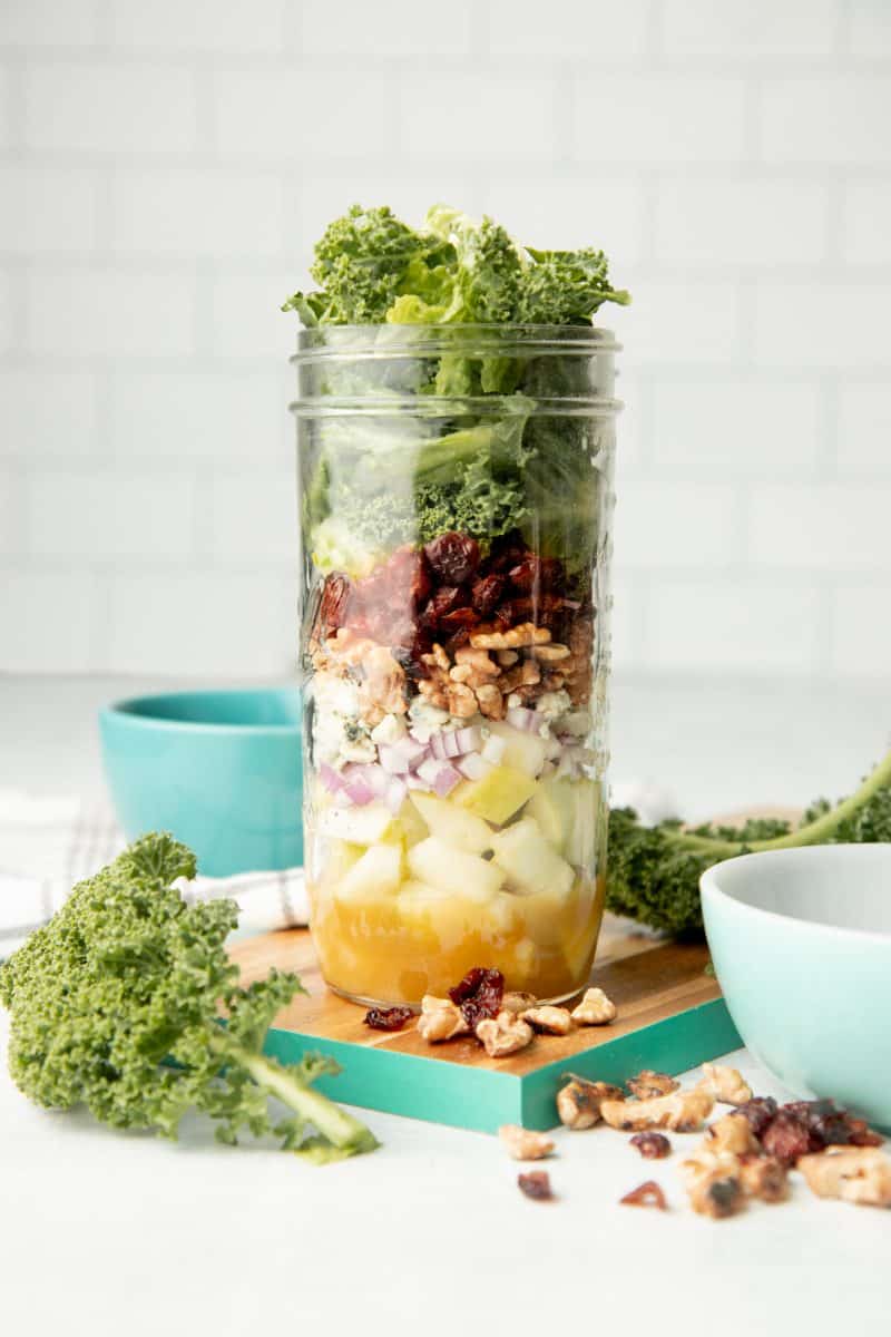 An apple walnut mason jar salad sits on a teal-edged cutting board, surrounded by kale and walnuts.