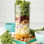 An apple walnut mason jar salad sits on a teal-edged cutting board, surrounded by kale and walnuts.