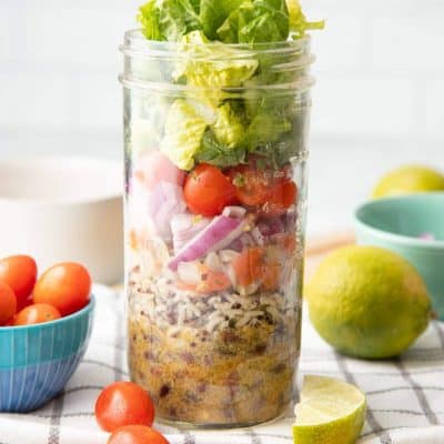 A glass jar layered with dressing, rice, beans, pico de gallo, onions, tomatoes, cilantro, and lettuce sits on a folded dish towel. Extra ingredients are scattered around the jar.