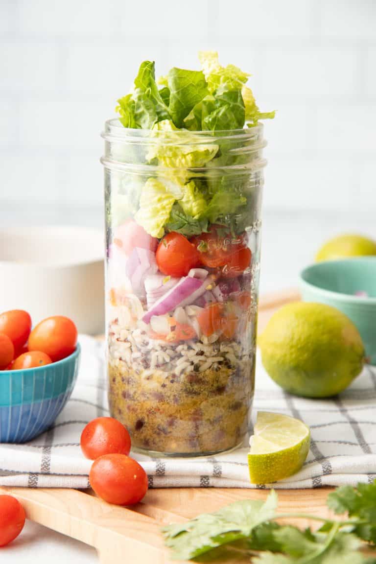 A glass jar layered with dressing, rice, beans, pico de gallo, onions, tomatoes, cilantro, and lettuce sits on a folded dish towel. Extra ingredients are scattered around the jar.