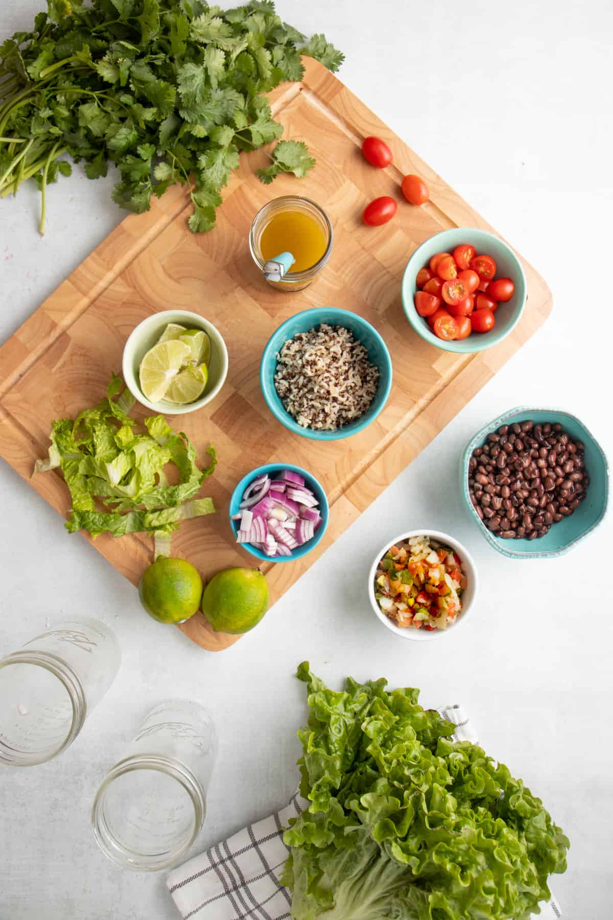 Ingredients for black bean fiesta salad are in blue bowls. Bowls are sitting on a wooden cutting board and a white countertop. Two empty jars and lettuce are on the counter.