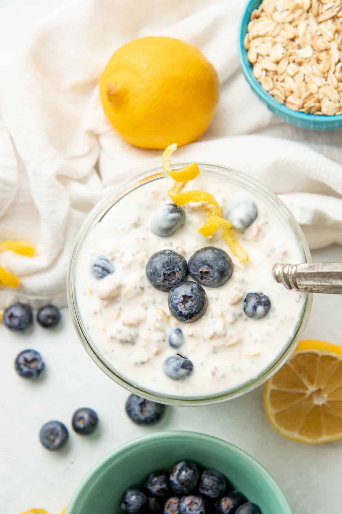 A spoon rests in a glass jar of blueberry overnight oats. Blueberries and a twist of lemon rest on top of the oatmeal.