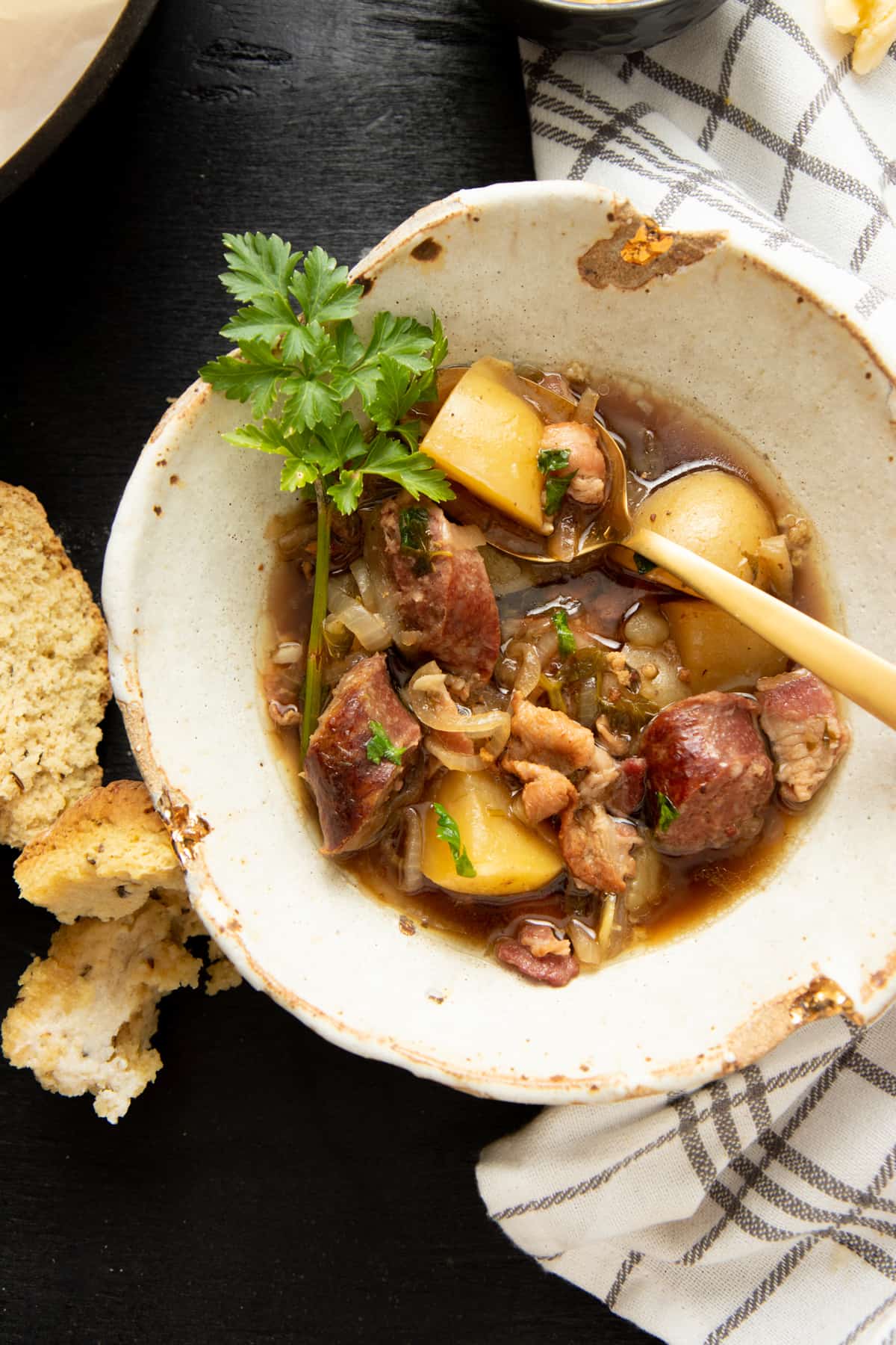 An ivory bowl is filled with hearty Irish stew. A gold spoon holds a potato and a piece of bacon. Soda bread is on the table.