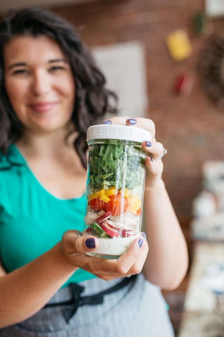 A smiling woman in a teal shirt and gray striped apron holds a tall mason jar. The jar is filled with layered salad ingredients, including dressing, tomatoes, peppers,and lettuce. The jar has a white lid.